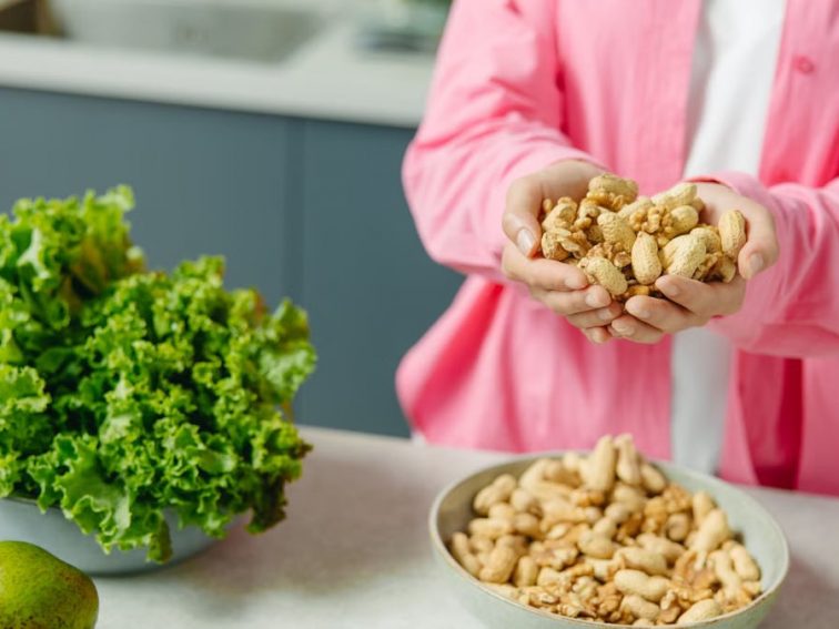 Hands presenting peanuts with a bowl, lettuce, avocado, and tomato nearby on kitchen counter.